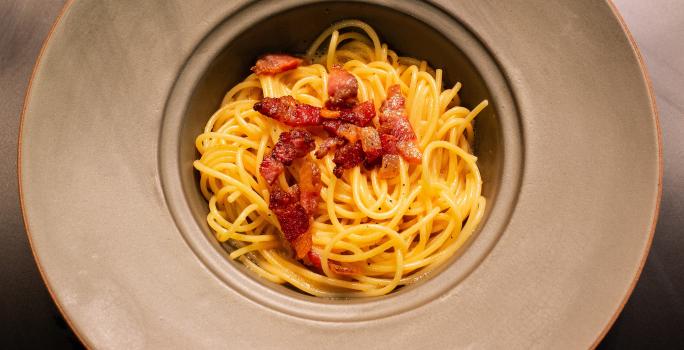 Fotografia em tons de amarelo com um prato cinza ao centro. Dentro do prato existe uma receita de macarrão carbonara finalizada com pedaços de guanciale frito.