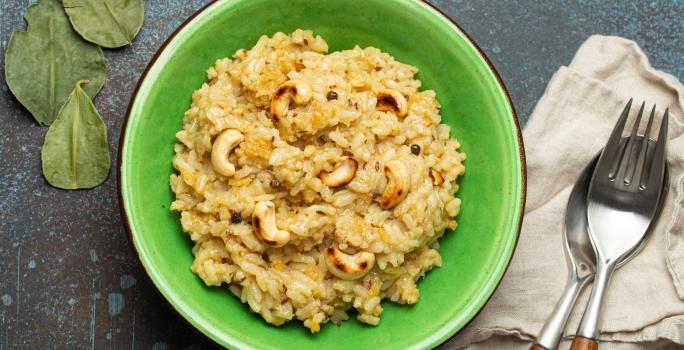 Fotografia vista de cima de um arroz crocante com castanha de caju dentro de um recipiente fundo verde, o qual está sobre uma mesa escura preta. Ao lado do recipiente tem um paninho e talheres por cima.