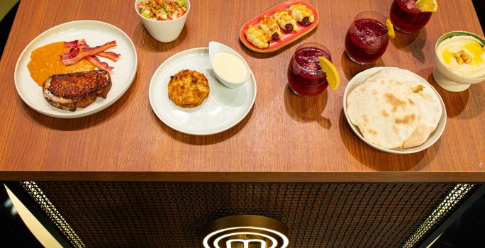 Fotografia de uma mesa de madeira com um brunch com pão sírio, hommus, ovos, frutas, massa folhada, creme inglês de requeijão, crumble de banana e torrada. Todas as partes estão porcionadas em recipientes brancos e separados.