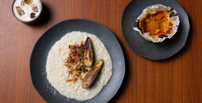 Fotografia vista de cima de três pratos pratos pretos com receitas diferentes, sendo elas: Fonduta de Gruyère com Cogumelos, Risotto de Brie com Abobrinha e Torta de Queijo com Compota de Frutas Amarelas. Os prato estão sobre uma bancada de madeira.