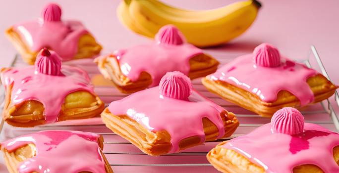 Fotografia de vários pasteis pequenos quadrados, recheados de banana e leite ninho, com cobertura de creme de nesquik. Os pasteis estão sobre uma grande branca, sobre uma mesa rosa.