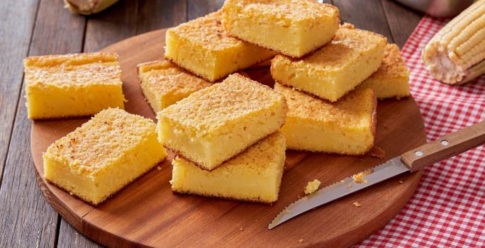 Foto da receita de bolo de milho cremoso, fatiado em pedaços sobre uma tábua de madeira sobre um pano quadriculado vermelho e branco. Na mesa há uma faca e espigas de milho