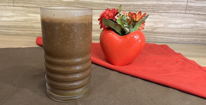 Fotografia em tons de marrom e vermelho com um copo vermelho ao centro. Dentro do copo existe uma bebida feita com suco de maracujá e achocolatado NESCAU, de cor marrom escura.