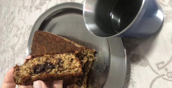 Fotografia de uma mão feminina segurando um pedaço de bolo de banana com aveia e chocolate. A outra parte do bolo está atrás da mão, sobre um recipiente oval de alumínio, ao lado de um copo de aluminio com café.