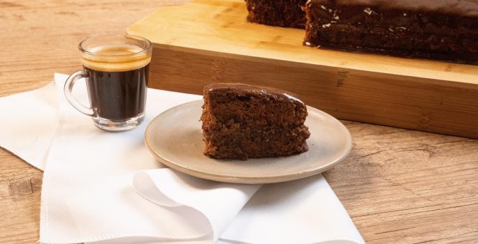 Fotografia de uma bancada de madeira, e sobre ela tem uma torta de chocolate com café em um prato de vidro em tom pastel, que está apoiado sobre um pano branco. Ao lado da sobremesa tem uma xícara com Nescafé, também sobre o pano.