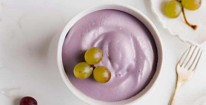 Fotografia em tons de branco e roxo com um pote branco ao centro. Dentro do pote existe uma receita de mousse de uva de cor roxa com algumas uvas verdes de enfeite.