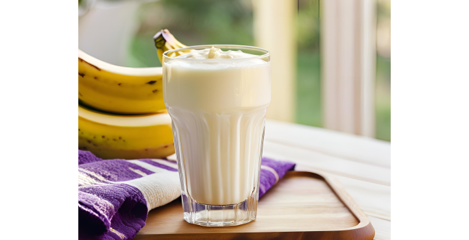 Fotografia em tons de roxo e amarelo com um copo de vidro ao centro. Dentro do copo existe um shake de coco, batata doce e banana de cor branca. Ao fundo existe duas bananas