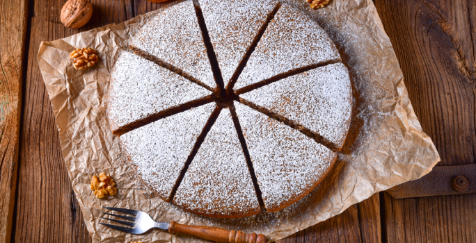 Foto em tons de marrom da receita de bolo alemão de mel servida cortada em 8 pedaços polvilhada com açúcar de confeiteiro, bolo em cima de uma mesa de madeira com nozes ao lado