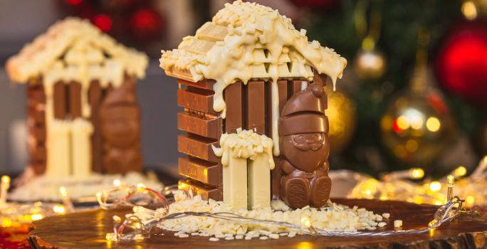 Imagem da receita de Casinha de KitKat Papai Noel, montada num formato miniatura de uma casinha de chocolate em tons marrom e branco, com um Papai Noel de chocolate à frente, sobre uma tábua de madeira e com decorações de natal ao redor