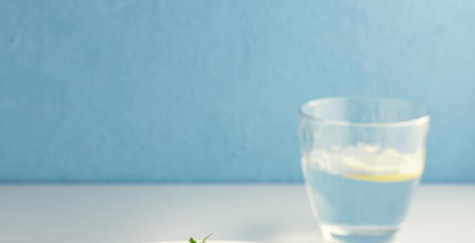 Fotografia de macarrão tipo parafuso com molho de tomate e berinjela dentro de um prato branco de vidro fundo. Ao redor da pasta tem um garfo e um copo de vidro com água e limão. O prato está sobre uma mesa branca.