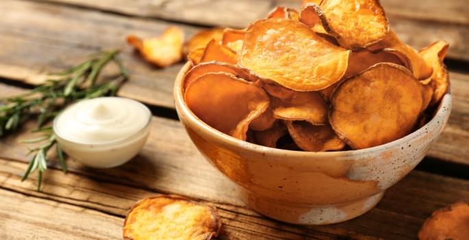 Fotografia em tons de marrom com um prato fundo de cerâmica ao centro. Em cima do prato existe uma porção de chips de batata doce feitos na Air Fryer