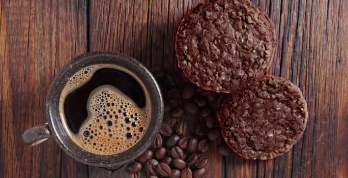 Foto vista de cima da receita de cookies de aveia e café, servidos em uma bancada de madeira, ao lado de uma xícara de café preparada com alguns grãos de café espalhados