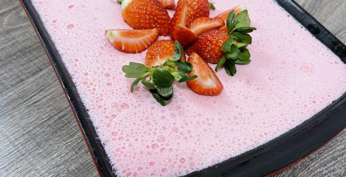 Imagem de uma receita de gelatina em tom rosa, decorada com morangos picados, servida em um refratário preto. Na imagem há um selo com o texto Receita do Consumidor em tom azul claro.