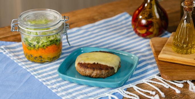 Fotografia em tons de azul com um prato azul ao centro. Em cima do prato existe uma receita de hambúrguer no prato acompanhada por uma fatia de queijo muçarela e uma salada no pote feita com alface e cenoura