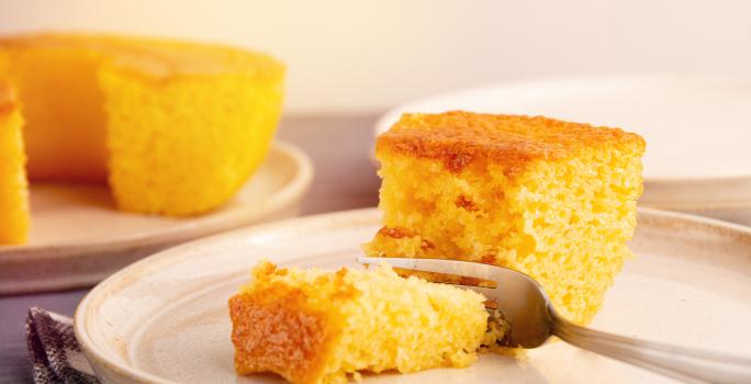 Fotografia de um prato grande de vidro com uma fatia grande de bolo de milho e um garfo cortando um pedaço. No fundo tem o bolo inteiro, sem essa fatia, sobre um apoio raso de cor clara.