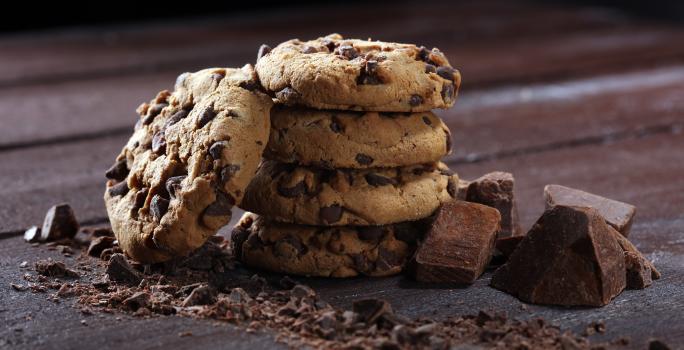 Fotografia de quatro cookies com gotas de chocolate, um sobre o outro. Encostado neles tem mais um cookie, ao lado de pedaços de chocolate, sobre uma mesa de madeira escura.