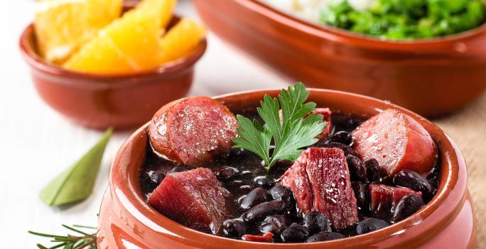 Fotografia de um recipiente médio marrom clássico com uma feijoada com caldo de bacon, linguiça e bacon com uma folha de salsinha. Ao fundo tem outros recipientes iguais, porém menores, com laranja, arroz, couve e farofa.