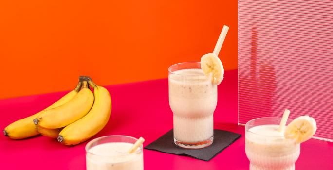 Fotografia de três copos de vidro com vitamina de banana e goiaba, e em cada copo, tem um canudo e uma rodela de banana decorando-o. Ao lado esquerdo, um cacho com quatro bananas, sobre uma mesa rosa com fundo laranja.
