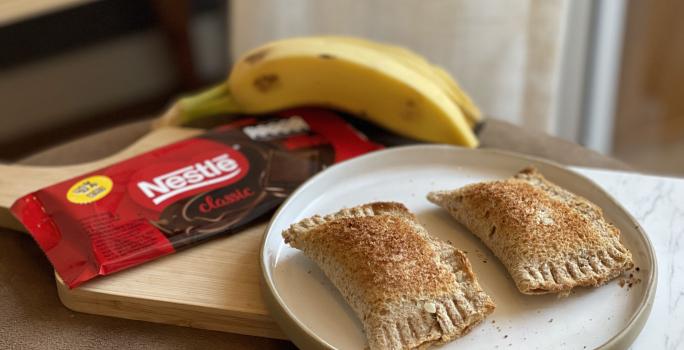 Fotografia em tons de marrom em uma bancada de madeira com uma tábua de madeira e ao centro, um prato redondo com dois pasteis. Ao lado, um tablete de chocolate e um cacho de bananas ao fundo.