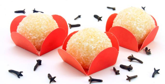 Fotografia de três unidades de beijinho com gelatina dentro de embalagens vermelhas sobre uma mesa branca, com cravos espalhados.