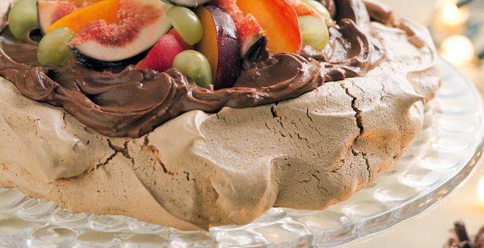 fotografia tirada de frente, contém um suporte para servir sobremesas transparente por cima um merengue coberto de chocolate com frutas para decorar.