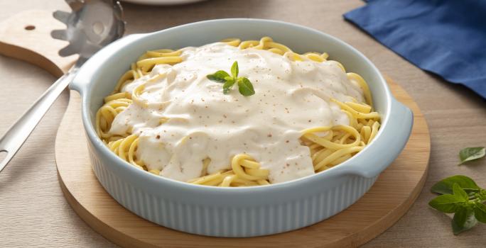 Fotografia em tons de azul em uma bancada de madeira, um pano azul, uma tábua de madeira redonda com um recipiente branco redondo com macarrão ao molho branco dentro dele. Ao lado, um pegador de macarrão.