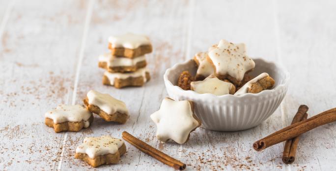 Fotografia de várias unidades de biscoito alemão em formato de estrela e uma pasta branca por cima. Uma parte dos biscoitos estão dentro de um pote de vidro branco, e o resto está espalhado em volta do refratário, sobre uma mesa de madeira branca.