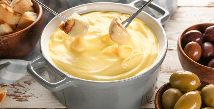 Fotografia em tons de branco com uma panela de fondue ao centro. Dentro da panela existe um fondue de queijo. Dois pedaços de pão estão sendo mergulhados no fondue de queijo. Ao fundo existe recipientes com azeitona e pedaços de pão.