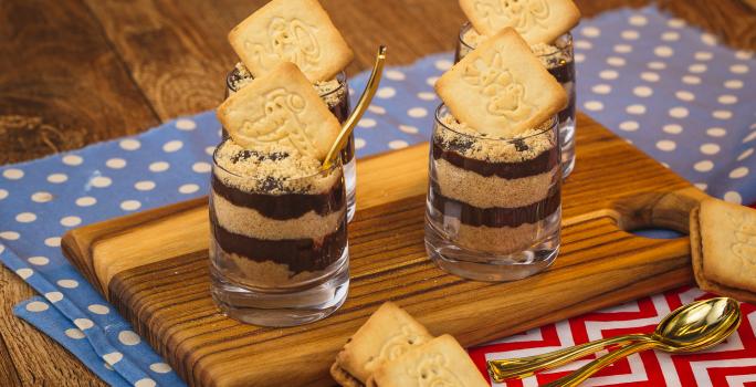 Imagem de uma bancada de madeira na qual há dois panos em tons azul claro e vermelho com texturas de bolinhas e listras. Há uma tábua pequena e sobre ela quatro copinhos com a receita em camadas de creme e biscoito triturado.