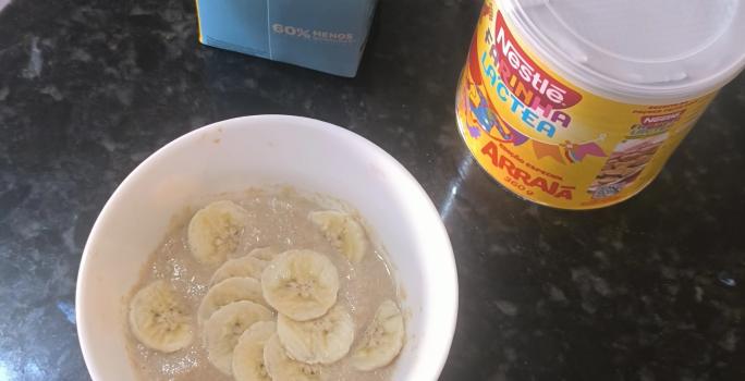 Fotografia de um recipiente branco fundo com um creme de farinha láctea com banana fatiada sobre uma bancada preta.