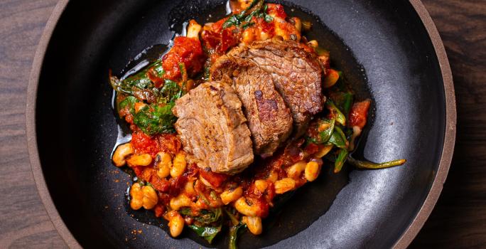 Fotografia de um prato grosso fundo preto com três fatias grossas de carne de sol sobre um molho de tomate e por baixo tem feijão manteiguinha e espinafre. O prato está sobre uma bancada de madeira.