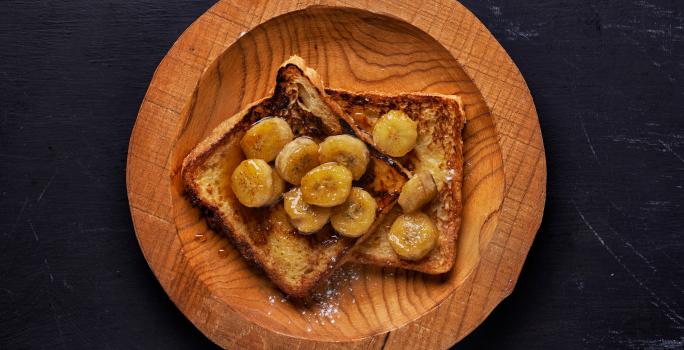 Fotografia em tons de madeira com um prato de madeira ao centro. Em cima do prato existe dois pães de forma cobertos com banana caramelizada e mel