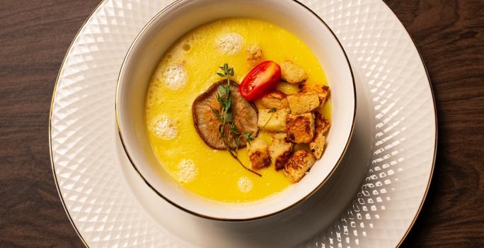 Fotografia em tons de marrom com um prato de sopa branco ao centro. Dentro do prato existe um creme de cor amarela feito com mandioquinhas acompanhado com croutons, cogumelos e tomate cereja.