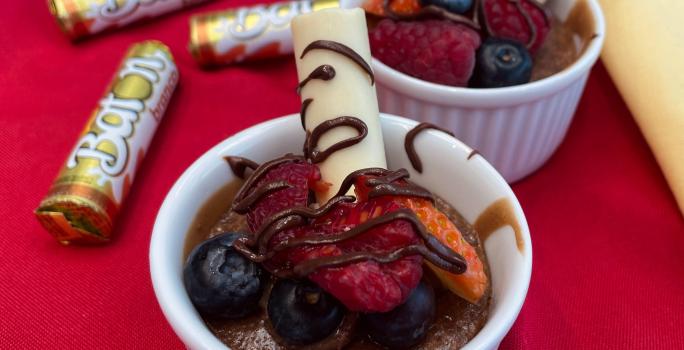 Fotografia em tons de vermelho com dois refratários ao centro. Dentro de cada um deles existe um bolo de chocolate cremoso coberto com morangos, calda de chocolate e BATON branco. Ao fundo existe embalagens de BATON branco.