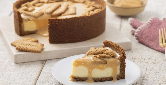 Foto da receita de Cheesecake de Tostines Especiarias. Observa-se uma torta com um pedaço tirado em um prato de sobremesa branco. A calda de doce de leite está escorrendo na fatia e biscoitos TOSTINES estão no topo decorando.