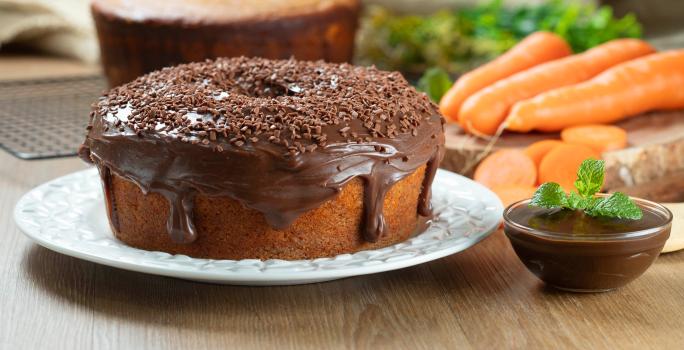 Fotografia de dois bolos de cenoura, um sem e o outro com cobertura de chocolate cremoso e granulado, apoiado em um prato branco. Ao lado tem mais cobertura em um recipiente pequeno de vidro, e ao fundo tem cenouras inteiras e alguns pedaços.