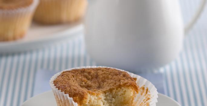 Fotografia em tons de azul em uma mesa de madeira com uma toalha listrada azul, um paninho azul, um pratinho de sobremesa branco com o muffin de banana com iogurte. Ao fundo, um bule branco com chá dentro.