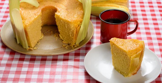 Foto de uma mesa com toalha vermelha quadriculada com dois pratos redondos, um com um bolo de fubá sem uma fatia, decorado com as folhas da espiga, e outro com uma fatia cortada em frente a uma xícara de café de louça vermelha