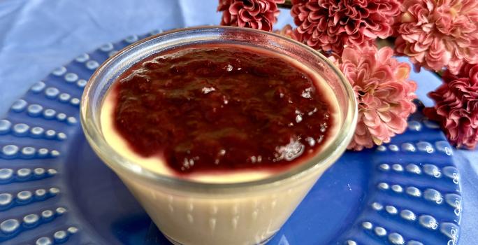 Fotografia em tons de azul e rosa com um pote transparente ao centro. Dentro do pote existe uma receita de flan de mamão coberto com geleia de frutas.