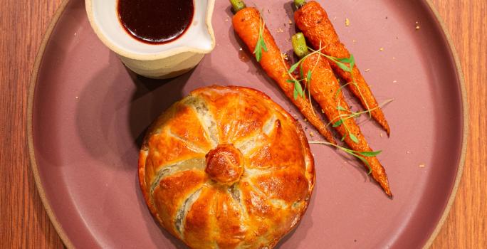 Fotografia em tons de marrom com um prato de cerâmica ao centro. Em cima do prato existe uma receita de Pithivier de Pato acompanhado de cenouras tostadas e molho de vinho do porto