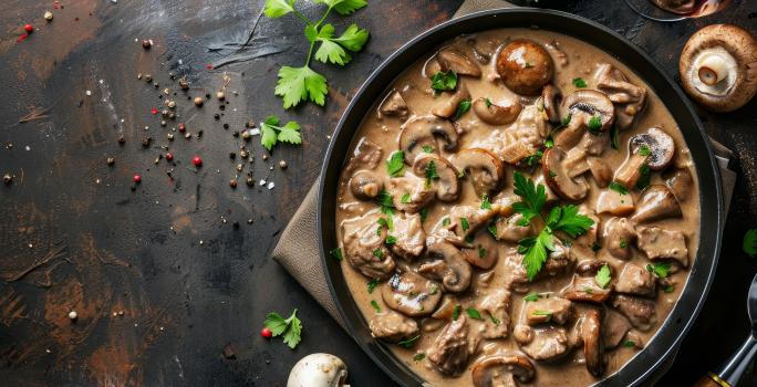 Fotografia vista de cima de uma frigideira de ferro com o molho madeira com champignon. Por cima tem algumas folhas de salsinha. A frigideira está sobre um pano marrom que está apoiado em uma mesa de madeira, com grãos de pimenta e folhas de salsinha