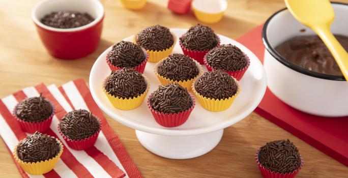 Fotografia em tons de vermelho e amarelo em uma bancada de madeira, um guardanapo de pano vermelho e branco listrado, uma tábua de madeira vermelha, um suporte para bolo branco com vários brigadeiros em forminhas amarelas e vermelhas em cima dele.