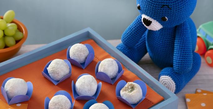 Fotografia em tons de branco em uma bancada de madeira de cor branca. Ao centro, uma mini mesa azul com uma toalha laranja em cima e dentro, há os docinhos. Ao lado, está o ursinho azul e ao fundo, há um carrinho e um pote de uvas verdes.