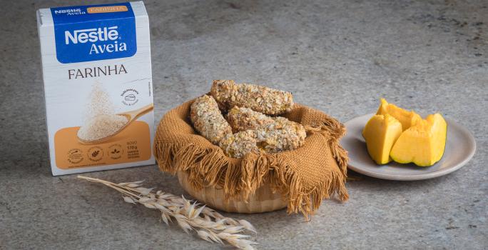 Foto receita de Croquete Vegetariano, servidos em uma cesta de vime, sobre uma bancada de mármore onde há uma caixa de nestlé farinha de aveia à esquerda e pedaços de abóbora sobre um prato à direita além de um ramo de trigo à frente