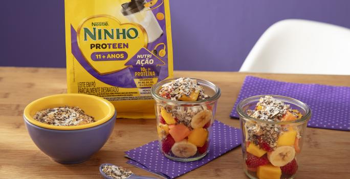 Fotografia de dois copos de vidro com frutas e granola sobre um paninho roxos com bolinhas brancas, sobre uma mesa de madeira. Ao lado dos copos, tem um recipiente amarelo e roxo com a granola, e tem uma embalagem de ninho proteen.