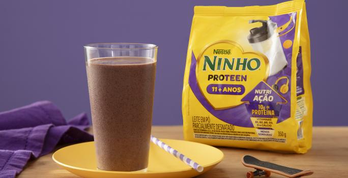 Fotografia de um copo de vidro grande com um smoothie de açaí apoiado em um prato amarelo raso com um canudo branco e roxo. Ao lado tem uma embalagem de ninho proteen, sobre uma mesa de madeira.