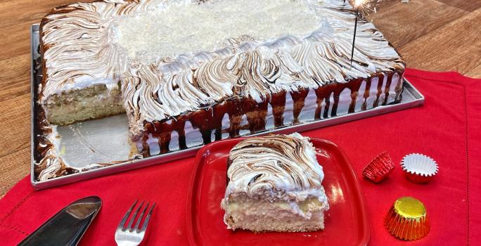 Foto da receita de Bolo de Aniversário de São Paulo, decorado com calda de chocolate, coco ralado. O bolo tem formato retangular e está sobre uma assadeira de metal, em uma bancada decorada com um tecido vermelho, sobre o qual há um prato com uma fatia