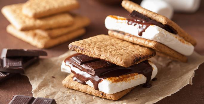 Fotografia de duas peças de s'mores sobre um papel manteiga. Ao fundo tem alguns pedaços de chocolate e alguns biscoitos empilhados, sobre uma mesa de madeira.