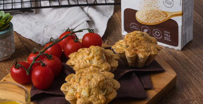 Fotografia em tons escuros de uma tábua de madeira quadrada com um pano marrom, tomates cereja e três tortinhas de legumes. Ao lado tem uma embalagem de farelo de aveia, e um pano branco com uma grade preta por cima, mais tortinhas apoiadas.