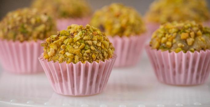 Fotografia de seis forminhas de cor rosa com brigadeiro de pistache enrolado no pistache triturado. Os brigadeiros estão em um apoio branco decorativo.